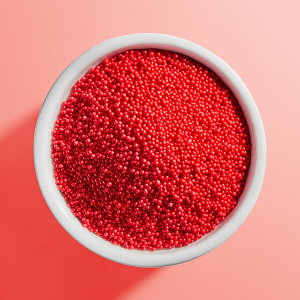 A white bowl filled with small, round, red candy sprinkles on a pink background.