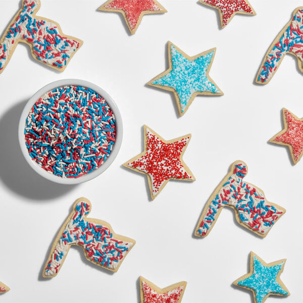 Star-shaped and flag-shaped cookies decorated with red, white, and blue sprinkles are arranged around a bowl filled with matching sprinkles.