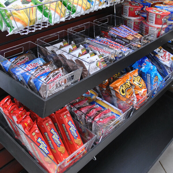 A set of clear plastic tote bins filled with assorted snacks and candy bars on a store shelf.