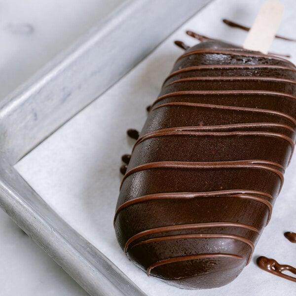 A chocolate-coated ice cream bar drizzled with additional chocolate, resting on a parchment-lined tray.