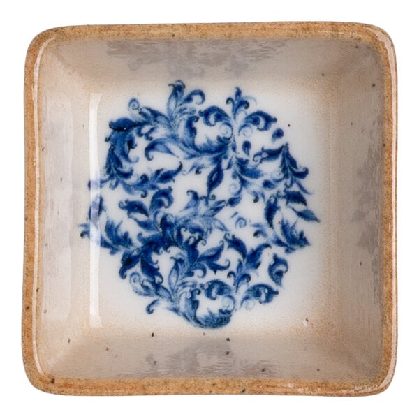 A square white porcelain ramekin with a blue floral design in the center and a tan rim.