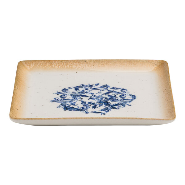 A square white porcelain plate with a blue floral design in the center and a tan rim.