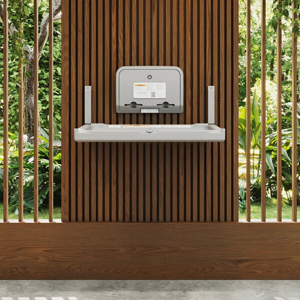A gray wall-mounted baby changing station with a dual liner dispenser installed on a wooden slat wall.