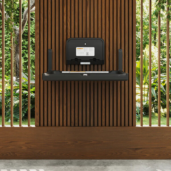 A black wall-mounted baby changing station with a dual liner dispenser, installed on a wooden slat wall.