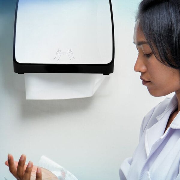 A person in a white coat is pulling a paper towel from a wall-mounted dispenser.