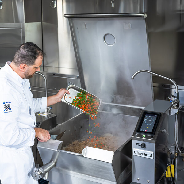 A chef is adding vegetables to a large Cleveland SGL-40-T1 PowerPan 40-gallon natural gas tilt skillet in a commercial kitchen.