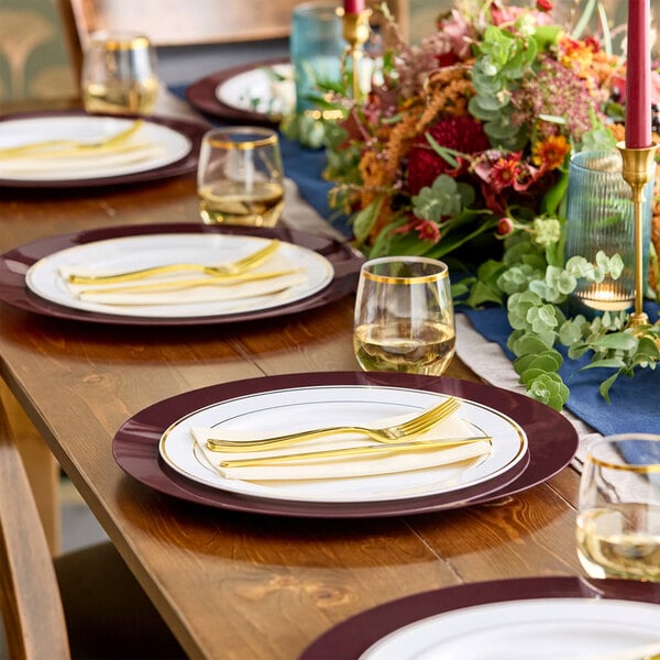 A wooden dining table is set with white plates featuring burgundy rims, gold utensils, clear glasses with gold rims, and a floral centerpiece with greenery and candles.