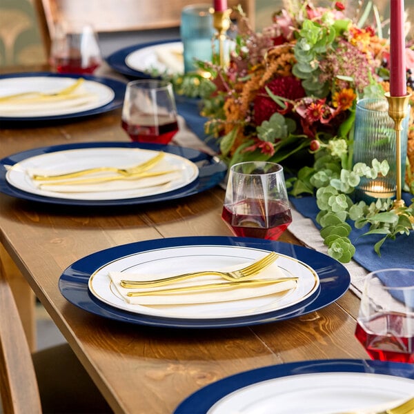 A wooden dining table is set with white plates featuring navy blue rims, gold-colored cutlery, glasses of red beverage, and a floral centerpiece with greenery and red flowers.