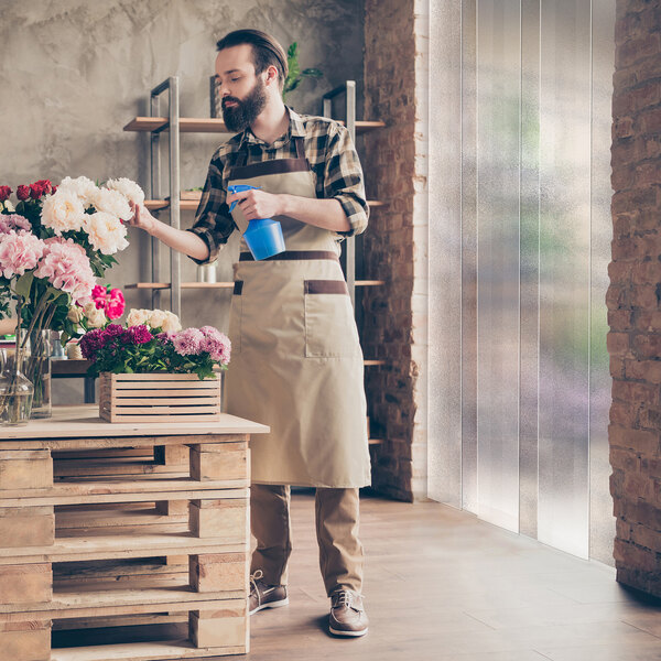 A person in an apron waters flowers in a shop with a frosted vinyl strip curtain hanging in the background.