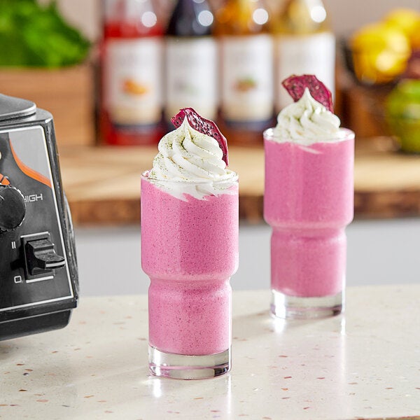 A pair of glasses filled with a bright pink smoothie topped with whipped cream and a slice of dried fruit, likely made from organic pitaya passion fruit smoothie cubes.