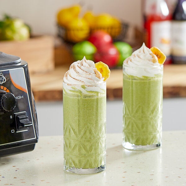 A pair of tall glasses filled with a green smoothie topped with whipped cream and banana slices, placed on a kitchen counter.
