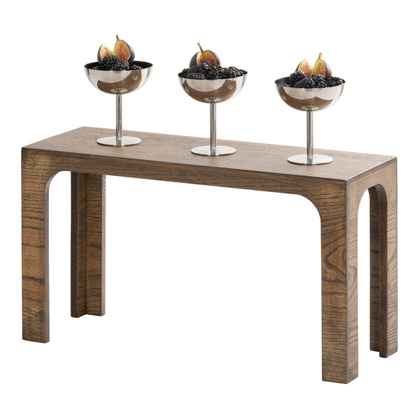 A rectangular chocolate brown oak wood riser with three metal bowls of fruit on top.