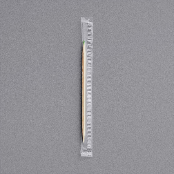 A single wooden toothpick with a green tip, individually wrapped in clear plastic, placed on a gray surface.