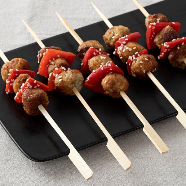 Several bamboo skewers with meatballs and red bell pepper pieces, garnished with sesame seeds, are arranged on a black rectangular plate.