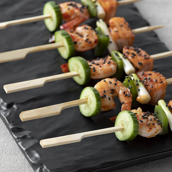 Several bamboo skewers with shrimp, cucumber slices, green peppers, and black sesame seeds are arranged on a black rectangular plate.