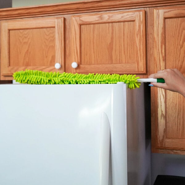 A green flexible microfiber wand duster being used to clean the top of a refrigerator.