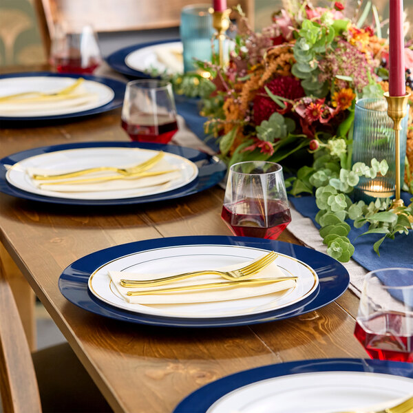 A wooden dining table is set with navy blue and white plates, gold-colored cutlery, glasses of red beverage, and a floral centerpiece with greenery and red flowers.