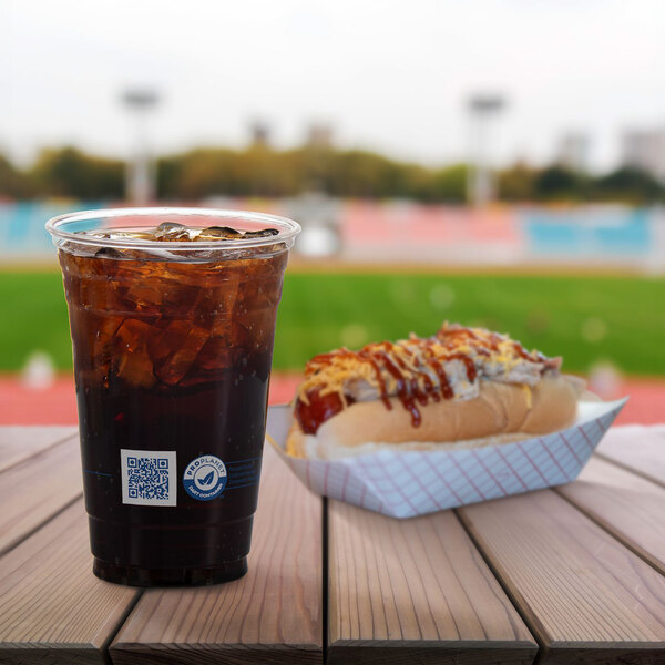 A clear 20 oz. plastic cold cup filled with a dark soda and ice, placed next to a hot dog in a paper tray on a wooden table.