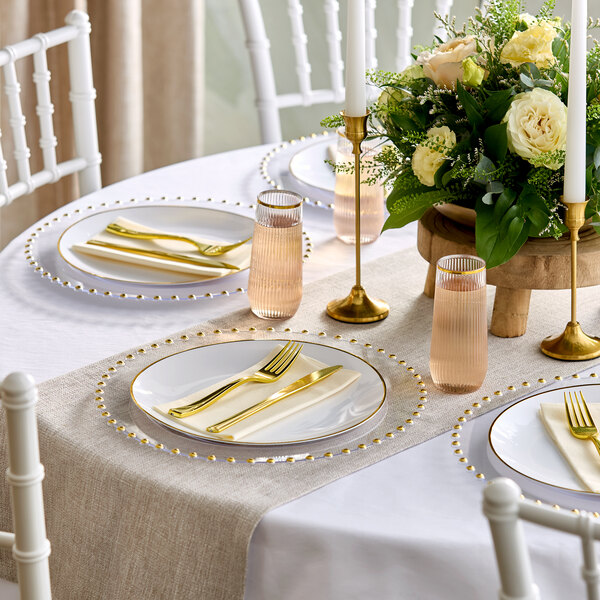 A round dining table is elegantly set with clear plates featuring gold beaded rims, gold-colored cutlery, ribbed glasses, white napkins, gold candle holders with white candles, and a floral centerpiece with white and cream flowers.