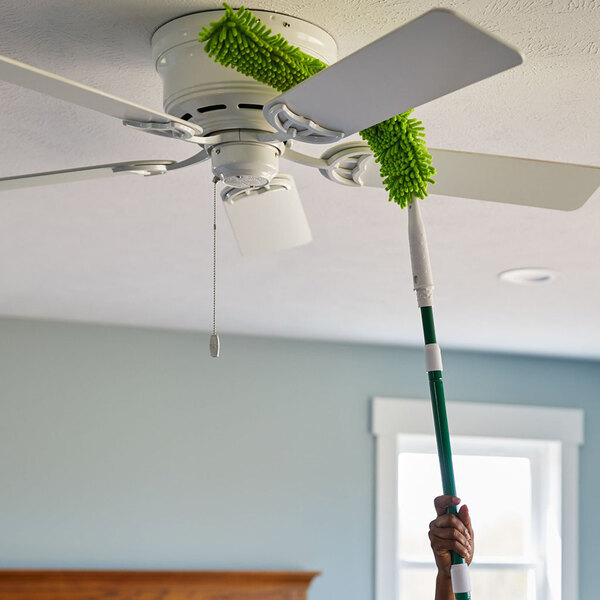 A green microfiber wand duster with an extendable handle being used to clean a ceiling fan blade.