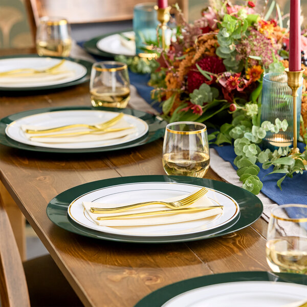 A wooden dining table is set with round plates featuring dark green rims, gold utensils, clear glasses with gold rims, and a colorful floral centerpiece.