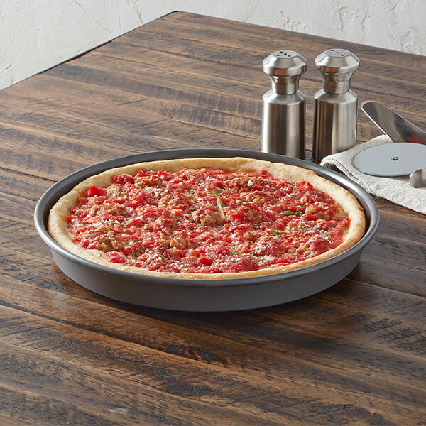 A round aluminum pizza pan with a deep edge, containing a baked pizza, placed on a wooden table.