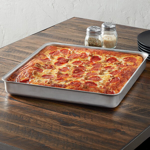 A square aluminum deep dish pizza pan containing a baked pizza, placed on a wooden table.