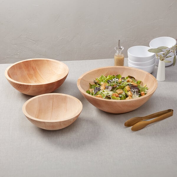 A large round maple wood serving bowl filled with salad, shown alongside two empty matching bowls.