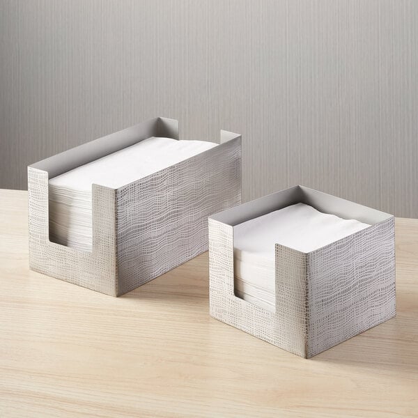 A square etched stainless steel napkin holder with a modern design, shown holding white napkins.