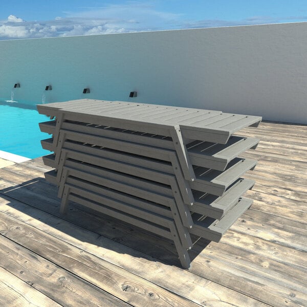 A stack of gray adjustable poly-lumber chaise lounges placed on a wooden deck near a pool.