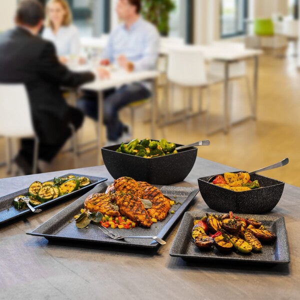 A square dark gray cast aluminum serving bowl filled with salad, shown on a table with other food dishes.