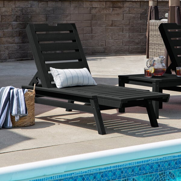 A black adjustable poly-lumber chaise lounge with a slatted design and a striped pillow, positioned beside a pool.