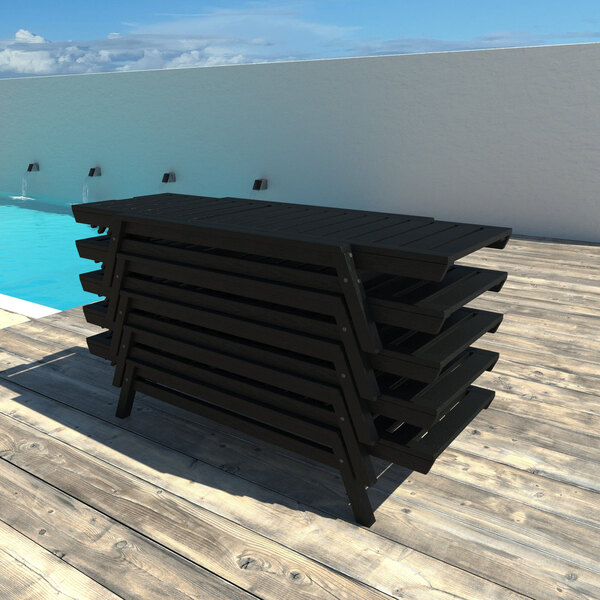 A stack of black adjustable poly-lumber chaise lounges on a wooden deck by a pool.