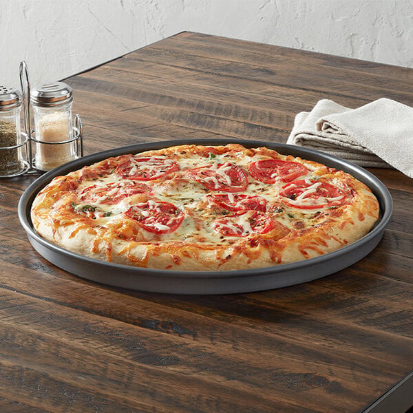 A round, straight-sided 14-inch aluminum pizza pan holding a baked pizza with tomato slices and cheese.