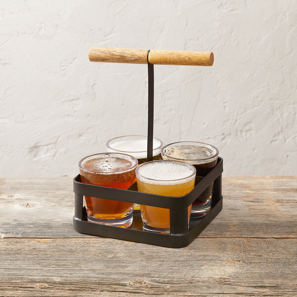 A black four-compartment tasting flight holder with a wooden handle, containing four glasses of different beverages.