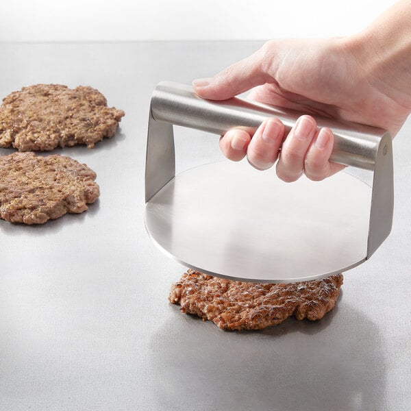 A round stainless steel burger press being used to flatten a hamburger patty on a flat surface.