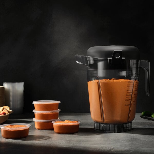 A large Vitamix blender with a 1.5 gallon container filled with an orange mixture, shown next to several small containers of the same mixture.