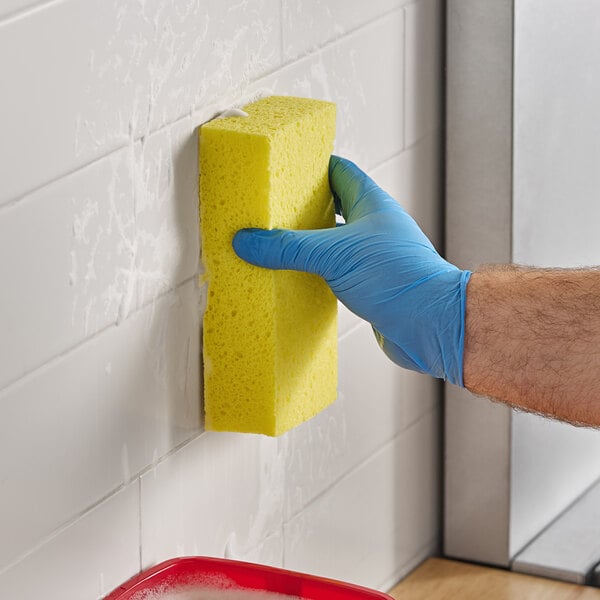 A person wearing a blue glove is cleaning a tiled wall with a large yellow sponge.