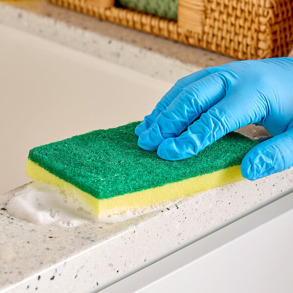 A hand wearing a blue glove is cleaning a countertop with a green and yellow sponge that has some soap suds.
