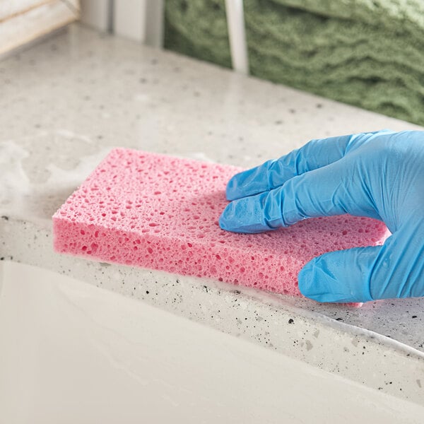 A hand wearing a blue glove is cleaning a speckled countertop with a pink rectangular sponge.