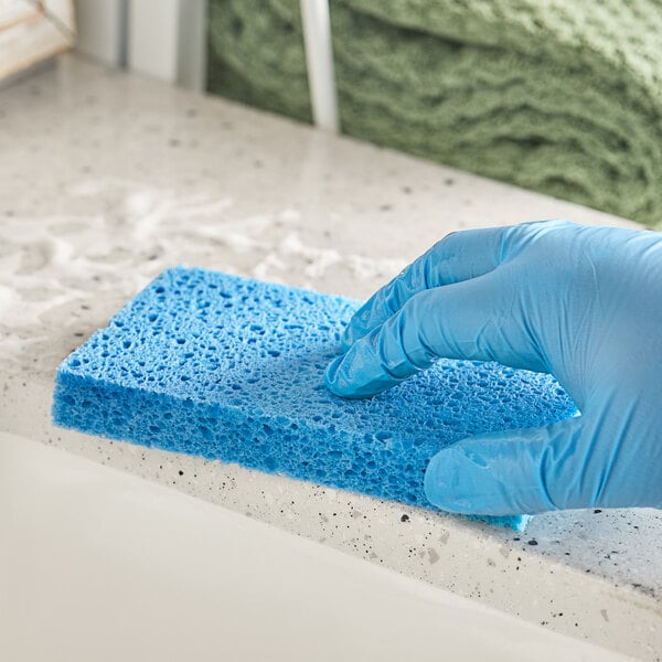 A gloved hand is cleaning a countertop with a blue rectangular sponge.