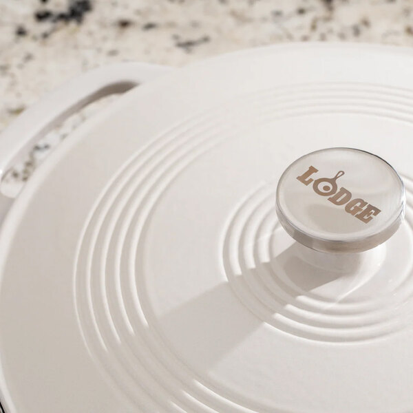 A white enameled cast iron casserole dish with a lid, featuring a metal knob labeled 'Lodge.'