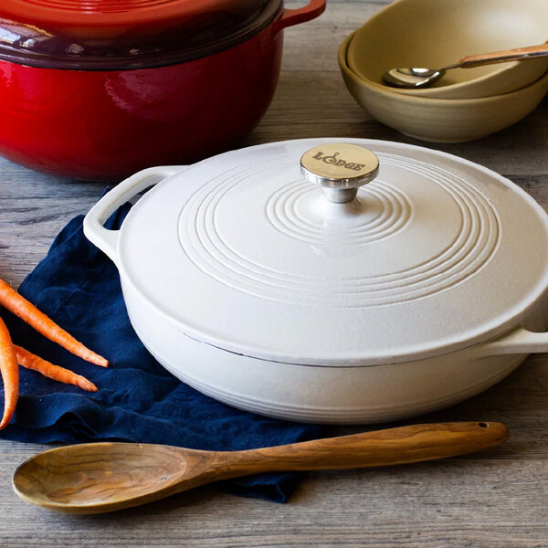 A white enameled cast iron casserole dish with a lid and handles, placed on a table with a wooden spoon and carrots nearby.