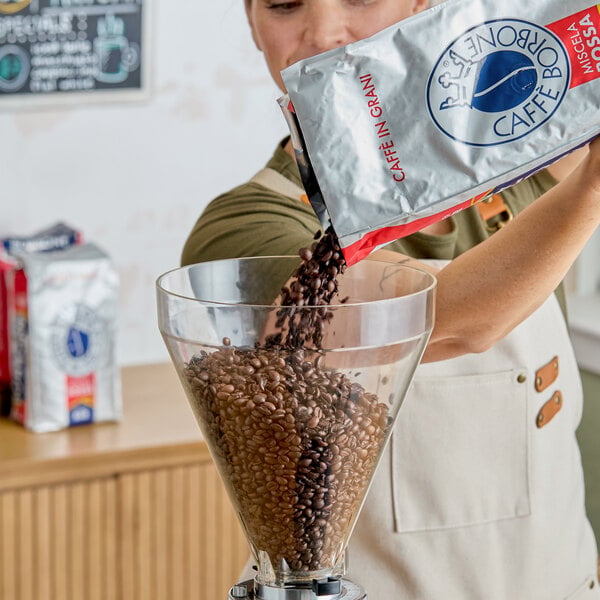 A person pours Caffe Borbone Red Blend whole bean espresso from a silver bag into a coffee grinder hopper.