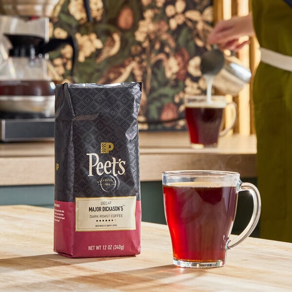 A bag of Peet's Coffee Decaf Major Dickason's Blend is placed on a table next to a clear glass mug filled with coffee, with another mug and a person pouring milk in the background.
