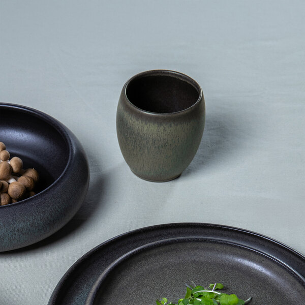 A small, rounded green and brown stoneware cup is placed on a light surface, surrounded by dark ceramic dishes.