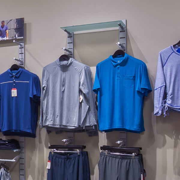 A vertical wall-mounted retail clothing display system with multiple hanging rails, faceouts, and glass shelves, showcasing various shirts and pants.