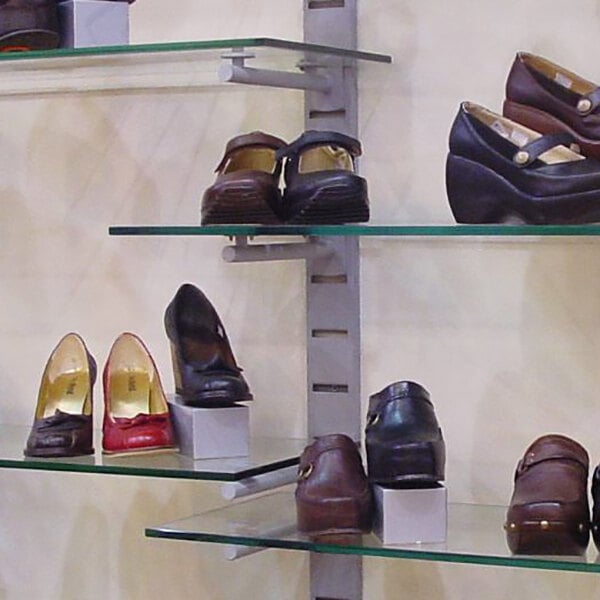 A vertical wall-mounted retail display with multiple glass shelves holding various pairs of women's shoes.