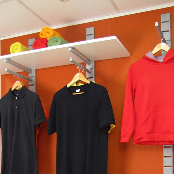 A vertical wall-mounted retail clothing display system with hanging rails, faceouts, and a shelf holding folded towels, showcasing shirts and a hoodie on hangers.