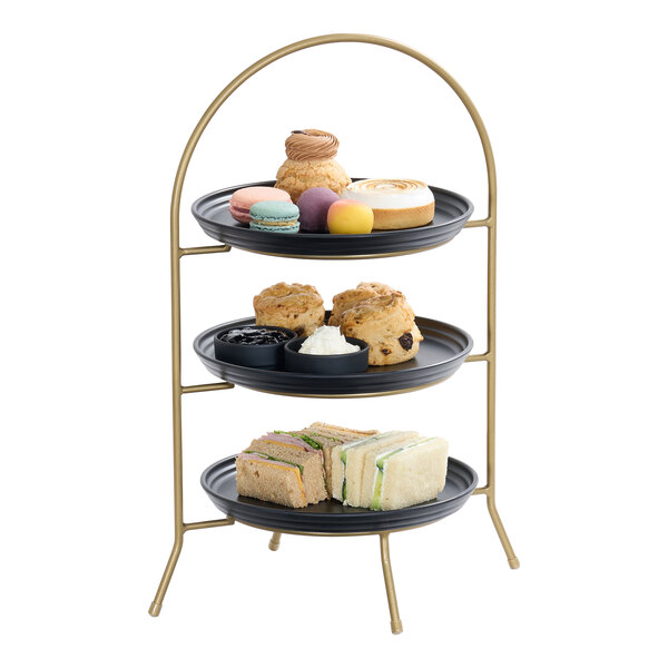 A three-tier matte gold plate stand displaying assorted pastries, scones, and finger sandwiches.